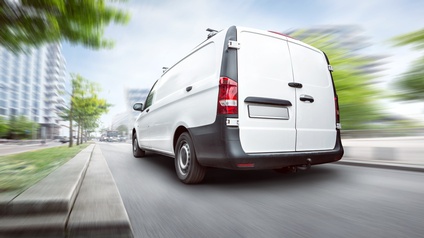 Ansicht des hinteren Hecks eines weißen Kleintransporters auf einer asphaltieren Straße. Links und rechts von der Straße sind grüne Bäume und weitere Gebäude bewegungsunscharf.