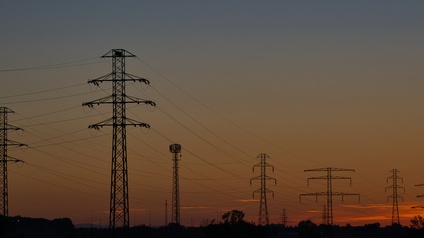 Aufnahme mehrerer versetzt stehender Strommasten, die durch Seile miteinander verbunden sind, im Sonnenuntergang.