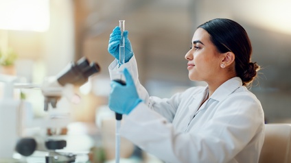 Seitliche Aufnahme einer Person in einem weißen Kittel, mit Handschuhen und geschlossenen Haaren, die eine große Pipette und ein Reagenzglas hält. Neben ihr steht ein Mikroskop