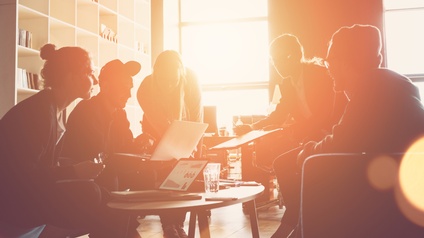 Gruppe von Personen sitzt rund um einen Tisch mit Arbeitsunterlagen und Laptops zusammen. Die Sonne scheint so durch das Fenster, dass man nur die Silhouetten der Personen sieht