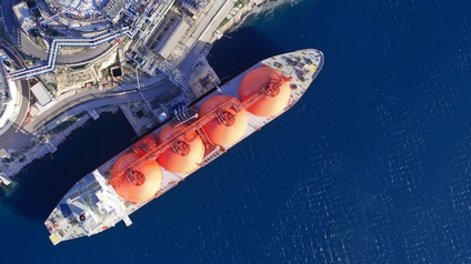 Top-Shot eines großen, langen Schiffes in einem blauen Gewässer. Auf dem Schiff sind vier große, orange Silos. Das Schiff ist direkt neben einer asphaltierten Oberfläche, auf der ein große Anlage steht.