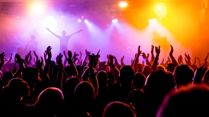 Rückenansicht einer Menschenmenge, in der teilweise Personen ihre Hände in die Luft gestreckt haben und klatschen. Ihnen gegenüber ist eine Bühne, auf der zwei Personen stehen. Die Bühne wird mit vielen Scheinwerfern beleuchtet