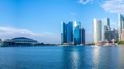 Skyline von Singapur, modere Wolkenkratzer, umrandet von Wasser und Bäumen unter blauem Himmel.