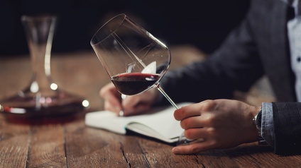 Eine Person hält mit der linken Hand ein Weinglas am Stiel, in dem sich etwas Rotwein befindet. In der rechten Hand hält die Person einen Stift, mit der sie etwas in einem gebundenem Heft notiert, das auf einem Holztisch liegt