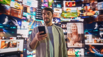 Mann blickt auf seinen Smartphonebildschirm. Hinter ihm ist ein bunter Hintergrund mit Bildschirmen zu sehen.