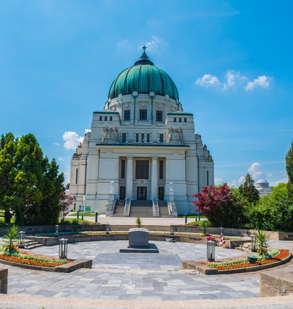 In der Mitte ist ein runder Platz mit einem steinern Sarg. Links und rechts davon sind grüne Büsche und Bäume. Neben dem Platz ist eine Aufbewahrungshalle mit einer grünen Kuppel