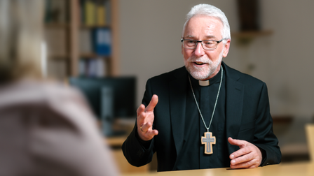 Ein älterer Mann in schwarzer Geistlichenkleidung mit weißem Kollar und Kreuzkette sitzt an einem Tisch und spricht gestikulierend mit einer Person gegenüber, die unscharf im Vordergrund zu sehen ist.