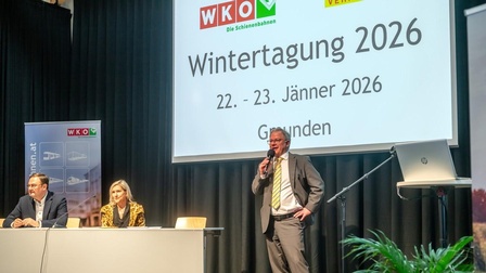 Eine Person in einem grauen Anzug steht vor einer Leinwand und hält ein Mikrofon. Auf der Leinwand steht Wintertagung 2026, 22.-23. Jänner Gmunden