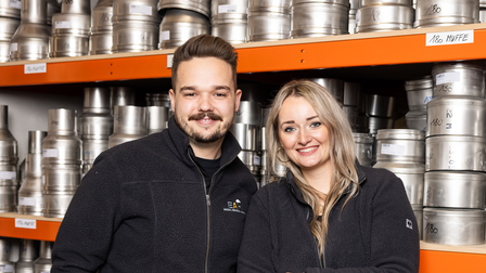 Lukas Steiner und Lisa Hupfauf stehen gemeinsam vor Regalen mit Edelstahlteilen in einem Lager. Beide tragen dunkle Firmenjacken mit EAH-Logo und lächeln in die Kamera. Im Hintergrund sind zahlreiche Metallteile und beschriftete Regalböden zu sehen.