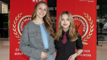 Zwei junge Frauen stehen vor einem roten Roll-up mit dem Logo des Junior Sales Champion. 
