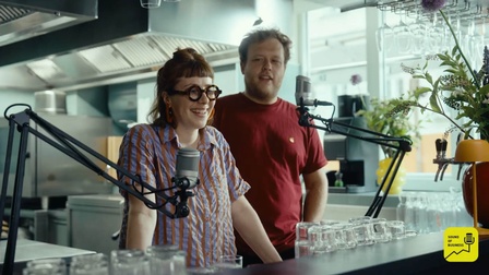 Frontale Aufnahme von zwei Personen, die hinter einem Tresen stehen. Vor den Personen sind zwei Mikrofone angebracht. Hinter ihnen ist eine Küche