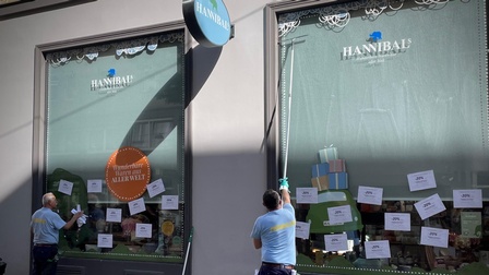 Vor zwei großen Fenstern steht jeweils eine Person. Die rechte Person fährt mit einem großen Fensterwischer über das Glas. Die rechte Person klebt daraufklebende Zettel ab. Auf den Fenstern steht unter anderem jeweils Hannibal