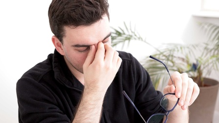 Mann mit geschlossenen Augen, in einer Hand hält er eine Brille, die andere Hand hält er zu seinen Augen