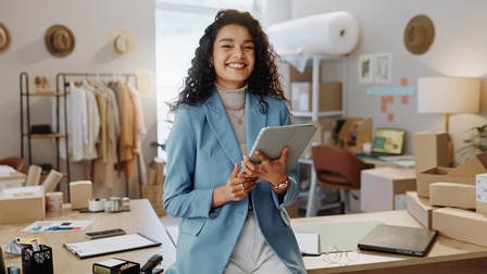Lächelnde Person mit Tablet in Händen lehnt an Tisch, im Hintergrund verschwommen Arbeitsutensilien, gestapelte Kartonagen, Gestell mit hängenden Gewändern, Regale und Arbeitsplatz mit Computer