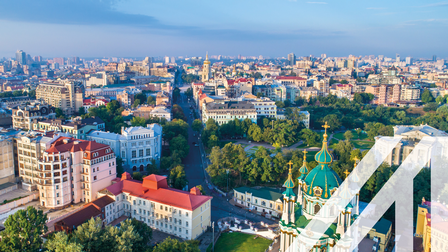 Stadtansicht von Kiew: eine breite Straße zieht sich durch die Stadt mit der Sankt-Andreas-Kirche und modernen Gebäuden, aufgelockert durch viele Bäume. Über das Bild wurde ein weißes Austria A gelegt.<br />
