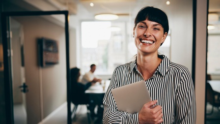 Porträt einer lächelnden Person mit dunklen Stirnfransen und gestreifter Bluse, die in einem Flur steht und mit beiden Händen einen Laptop beim Körper hält, im Hintergrund zeigen sich helle Meetingräume