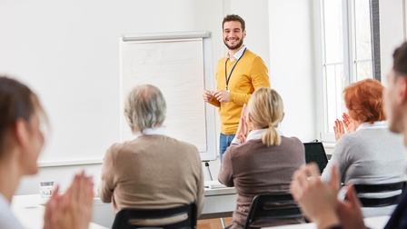 Person steht vor Flipchart und lächelt applaudierendes Publikum an