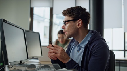 Seitliche Aufnahme einer Person mit kurzen, braunen Haaren und Brille, die an einem Schreibtisch im Büro sitzt. Vor ihr sind zwei Bildschirme. Die Person blickt auf den linken Bildschirm