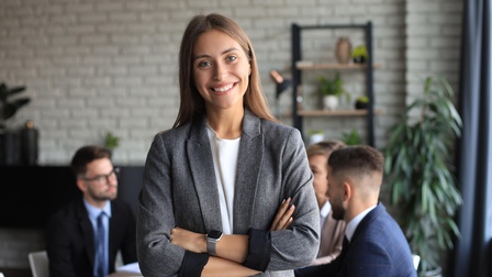 Lächelnde Person mit verschränkten Armen in Besprechungszimmer, im Hintergrund weitere Personen an Tisch sitzend