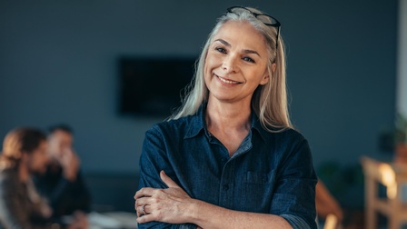 Person mittleren Alters mit langen weiß-grauen Haaren, Brille und blauem Jeanshemd steht lächelnd und mit verschränkten Armen in einem Raum während im Hintergrund andere Personen sitzen