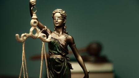 Statue Justitia, im Hintergrund sind Bücher sowie ein Richterhammer in der Unschärfe platziert