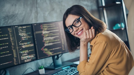 Lächelnde Person mit Brillen stützt Kopf in die Hand an Schreibtisch mit Monitoren, auf denen Quellcodes zu sehen sind, sitzend
