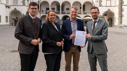 Drei Männer und eine Frau stehen im Landhaushof. Das Landhaus ist im Hintergrund zu sehen. Ein Mann übergibt ein Papier an einen anderen. 