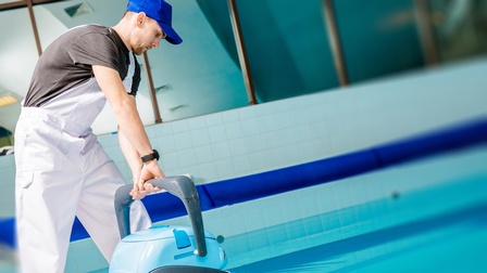 Eine Person mit heller Latzhose und Kappe steht neben dem Schwimmbecken in einem Hallenbad und gibt ein automatisches Reinigungsgerät in das Wasser.
