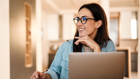 Person mit Brille sitzt vor einem Laptop mit Notizbücher und Stift an einem Tisch und blickt fröhlich und interessiert auf die Seite