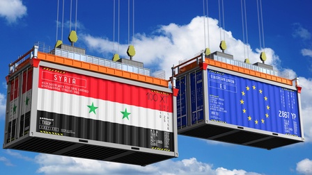 Zwei von Seilen hochgezogene Warencontainer: einer mit der Flagge Syriens, einer mit der EU-Flagge