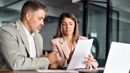 Geschäftsleute mit Dokumenten in der Hand im Büro, sitzend an einem Tisch vor einem offenen Laptop