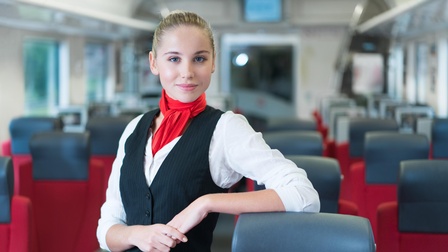 Person mit rotem Halstuch, weißem Hemd und schwarzem Gilet steht in Gang eines Wagons und stützt sich an einem Sitz ab