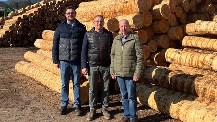 Drei Personen stehen im Freien vor großen, gestapelten Baumstämmen auf einem Holzlagerplatz, im Hintergrund Wald und blauer Himmel.