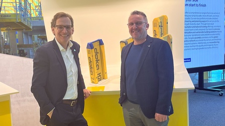 Zwei Personen stehen an einem gelben Messestand mit der Aufschrift „Formwork beams | Schalungsträger“, im Hintergrund eine Messehalle mit Gerüsten und Monitor.