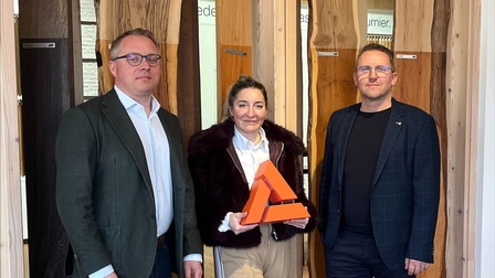 Drei Personen stehen in einem Innenraum vor einer Präsentationswand mit verschiedenen Holzoberflächen; eine Person hält eine dreieckige, orangefarbene Auszeichnung in den Händen.