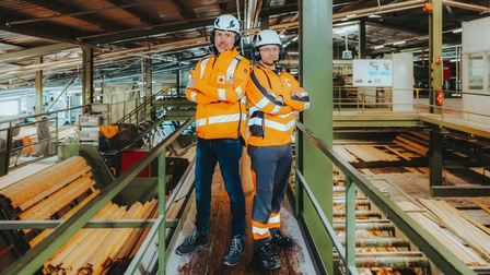 Zwei Personen in orangefarbener Schutzkleidung und Helmen stehen auf einem Steg in einer industriellen Holzverarbeitungshalle mit Förderanlagen und gestapeltem Schnittholz.