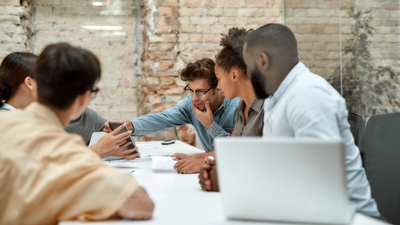 Sechs Personen sitzen sich an einem langen, hellen Tisch in einem Raum mit Glaswänden gegenüber. Eine Person links zeigt den rechts sitzenden Person etwas auf einem Laptop