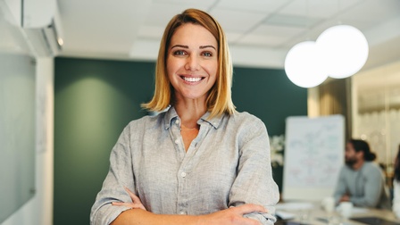Halbporträt einer Person in einem grauen Hemd und mit verschränkten Armen in einem Raum stehend. Hinter ihr ist ein langer Tisch, an dem zwei Personen sitzen sowie ein Flipchart. Auf dem Tisch liegen Unterlagen.