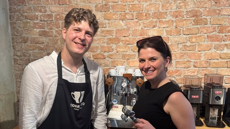 Zwei Personen stehen vor einer Espressomaschine und Kaffeemühlen in einer Kaffeerösterei oder Barista-Schulung. Eine Person trägt eine Schürze mit Corretto-Coffee-Logo, die andere hält einen Siebträger in der Hand. Im Hintergrund ist eine Backsteinwand zu sehen.
