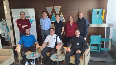 Gruppe von sieben Personen in einem Firmenfoyer, einige sitzen auf Sesseln, andere stehen dahinter. Im Hintergrund sind ein Firmenlogo 