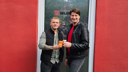 Stephan Pichler-Holzer überreicht Cornel Istrate das orange JW Häferl vor einer Eingangstür.