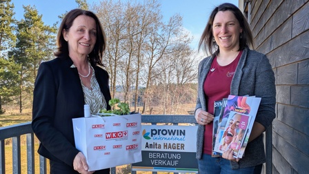 Fiw Obfrau Anne Blauensteiner und die Unternehmerin Anita Hager stehen auf einer Terrasse vor Frau Hagers Firmen­schild und halten eine WKO‑Geschenktasche sowie Informationsbroschüren. Im Hintergrund sind Bäume und eine Holzfassade zu sehen.