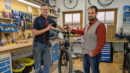 Zwei Personen stehen in einer Fahrradwerkstatt neben einem Mountainbike. Im Hintergrund sind Werkbänke mit Werkzeugen, Ersatzteilen und Regalen sichtbar.