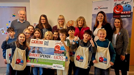 Gruppenfoto von mehreren Kindern und Erwachsenen in einem Innenraum. Die Kinder stehen vorne und halten  einen großen Spendenscheck über 1.500 Euro sowie Stoffbeutel mit dem Logo der Aktion der Direktberater. Hinter ihnen stehen mehrere Erwachsene. Rechts im Bild sind zwei Roll-up-Banner der Aktion 