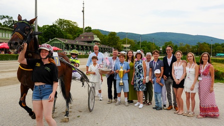 Siegerehrung auf einer Trabrennbahn: Ein Trabrennpferd mit Sulky steht neben einer Gruppe von Erwachsenen und Kindern, die einen Pokal und Geschenkkorb präsentieren. Im Hintergrund sind die Rennbahntribüne und bewaldete Hügel zu sehen.
