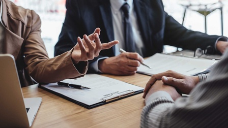  3 Personen sitzen an einem Tisch und führen eine Verhandlung. Unterlagen sind zu sehen, die Personen sind nur im Rumpfbereich zu sehen.