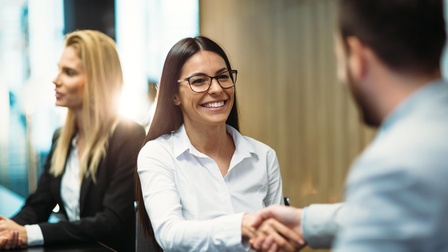Person mit dunklen langen Haaren und Brille reicht einer anderen Person freudig die Hand