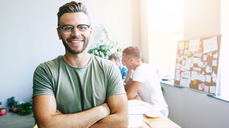 Porträt von einer lächelnden Person mit Brille, kurzen Haaren und T-Shirt mit verschränkten Armen, im Hintergrund sitzen weitere Personen an einem Tisch sowie steht eine Pinnwand mit Notizen am Fensterbrett