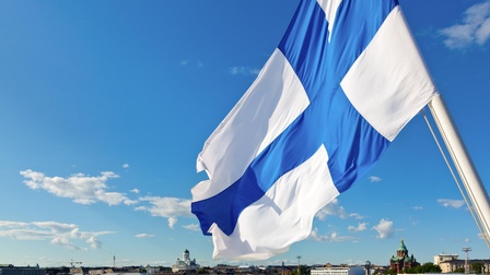 Die finnische Flagge weht im Wind, im Hintergrund eine Stadtansicht.