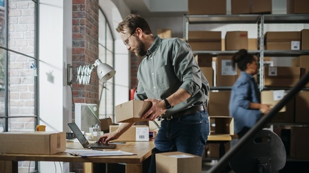Personen arbeiten im Lagerraum am Laptop, bereiten Pakete für die Auslieferung vor und verpacken sie.